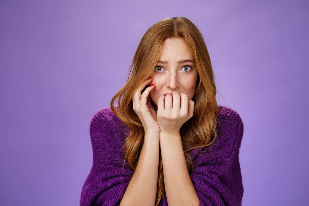 Scared Insecure Woman with Red Hair Biting Fingernails Feeling Frightened as Home Alone Hearing Weird Horrifying Noises Shaking from Fear Standing Anxious and Worried over Purple Background